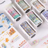 A collection of decorative transparent washi tape rolls in various floral and patterned colors displayed in cardboard boxes.