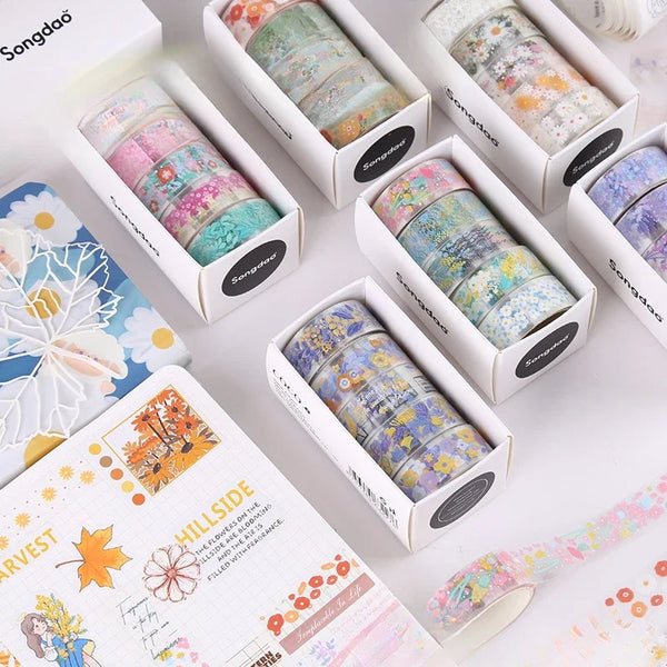 A collection of decorative transparent washi tape rolls in various floral and patterned colors displayed in cardboard boxes.