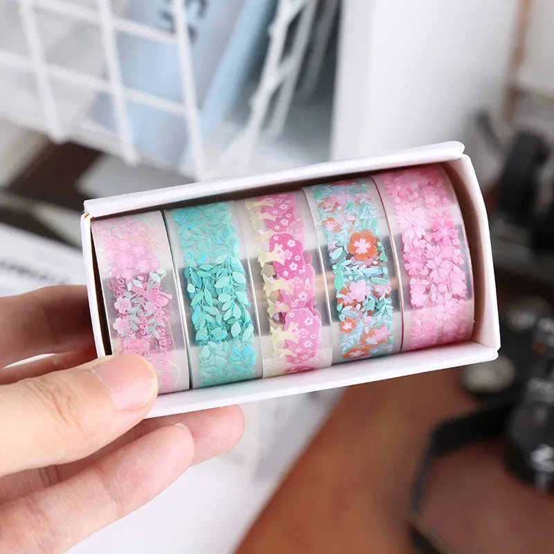 A hand holds a white box containing a set of decorative transparent washi tape rolls in various shades of pink and teal.