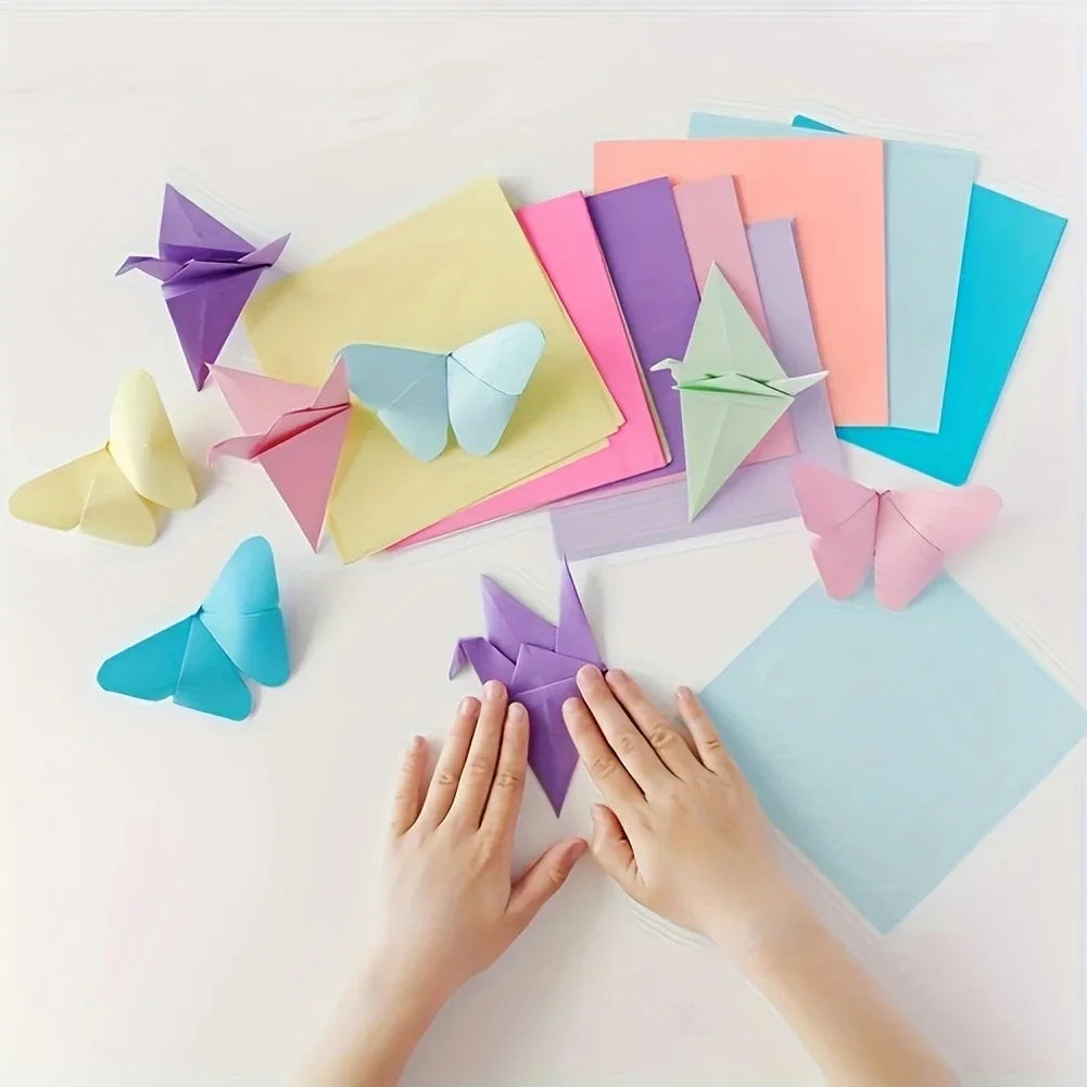 Hands are folding colorful cardstock sheets into origami shapes on a white surface.