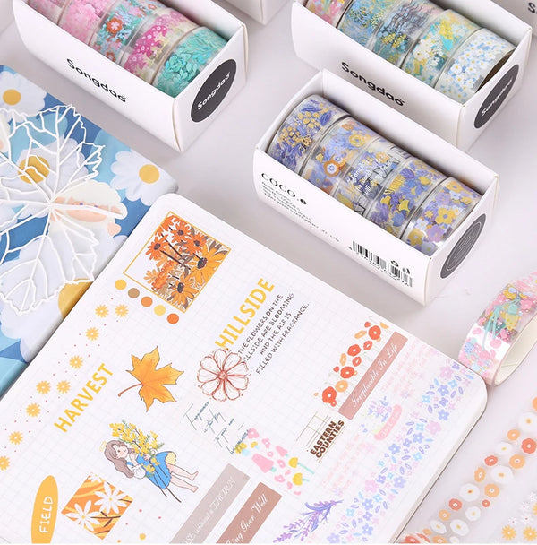 A collection of decorative transparent washi tape rolls in various colors and patterns displayed in open boxes next to a note