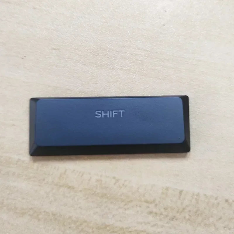 A black rectangular keycap labeled SHIFT resting on a light wooden surface.