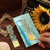 A vintage artistic metal bookmark with a turquoise tassel is displayed on dark wood next to a sunflower.