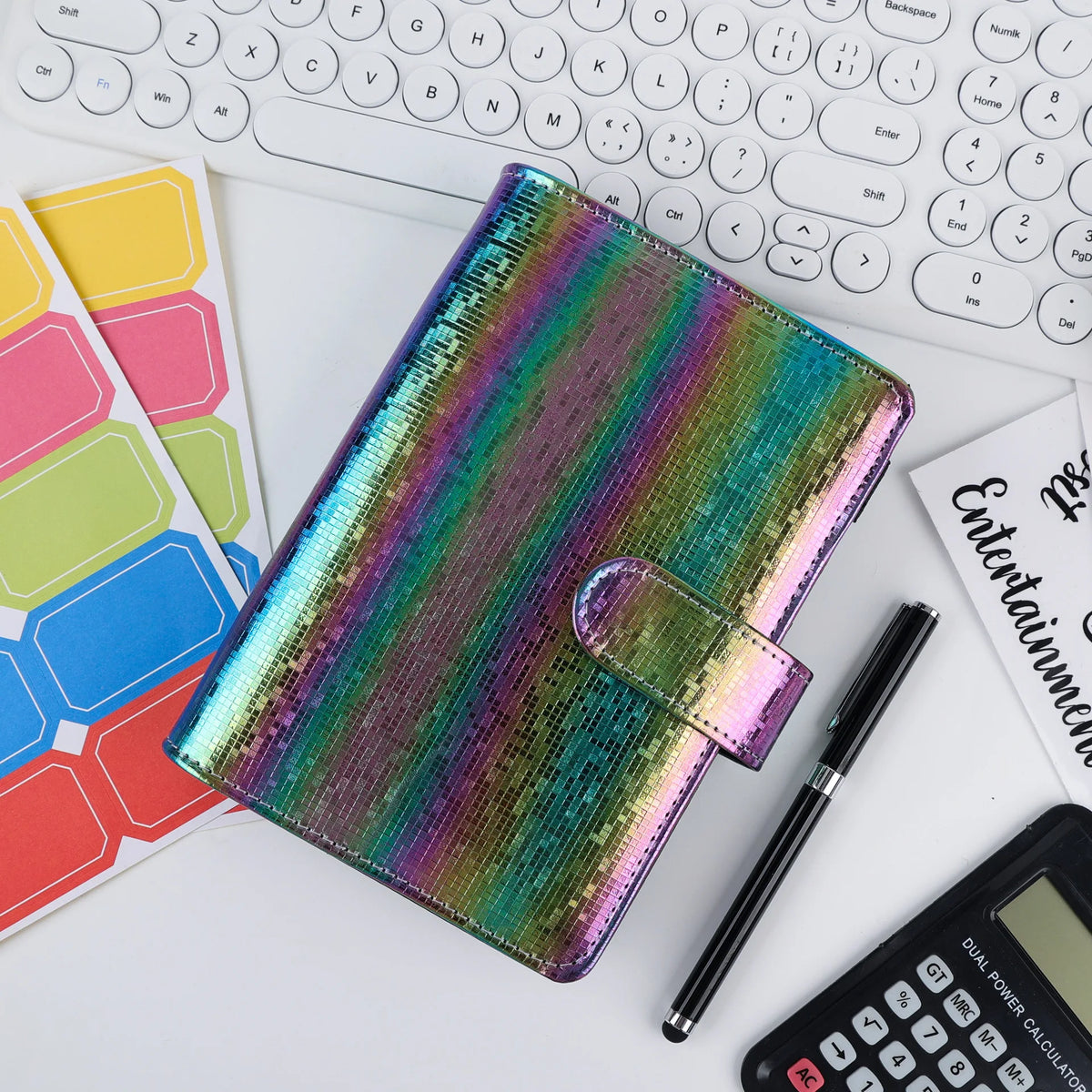 Un set de cahier et de stylo coloré avec des paillettes posé sur un bureau blanc à côté du clavier d'un ordinateur portable.