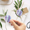 Hand placing a purple thank you sticker on white paper tied with natural twine and green leaves.