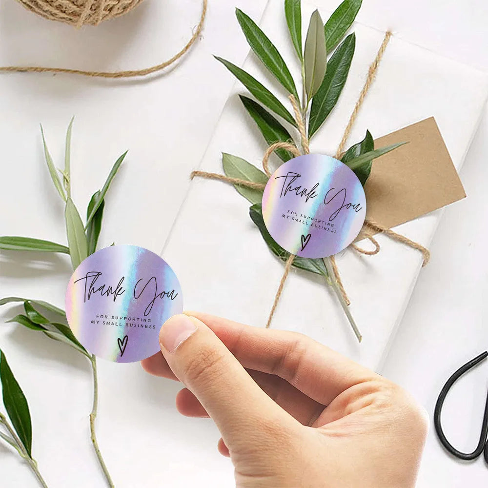 Hand placing a purple thank you sticker on white paper tied with natural twine and green leaves.