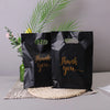 Two black thank you gift bags with gold script text are displayed next to green foliage.