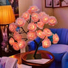 A pink rose tree LED night light displayed on a wooden table next to a blue armchair.