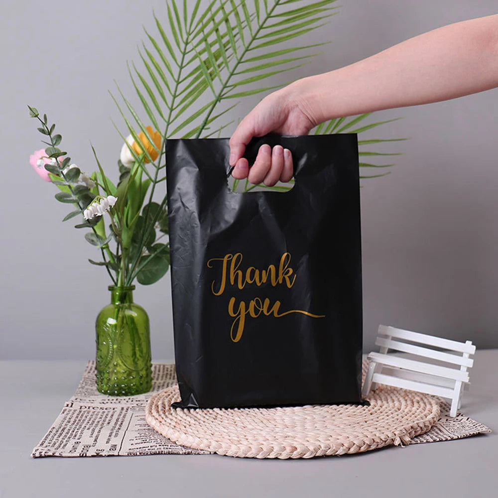A hand holds a black thank you gift bag with a gold script, resting on a woven mat next to green foliage.