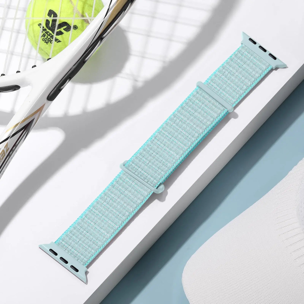 A light blue adjustable sport hook and loop watch strap is shown next to a yellow sports ball.