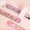 A pink plastic measuring tool with cat paw markings is shown next to a ruler and a white ribbon.