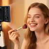 A woman applies makeup using a brush in front of a portable clip-on LED selfie light.
