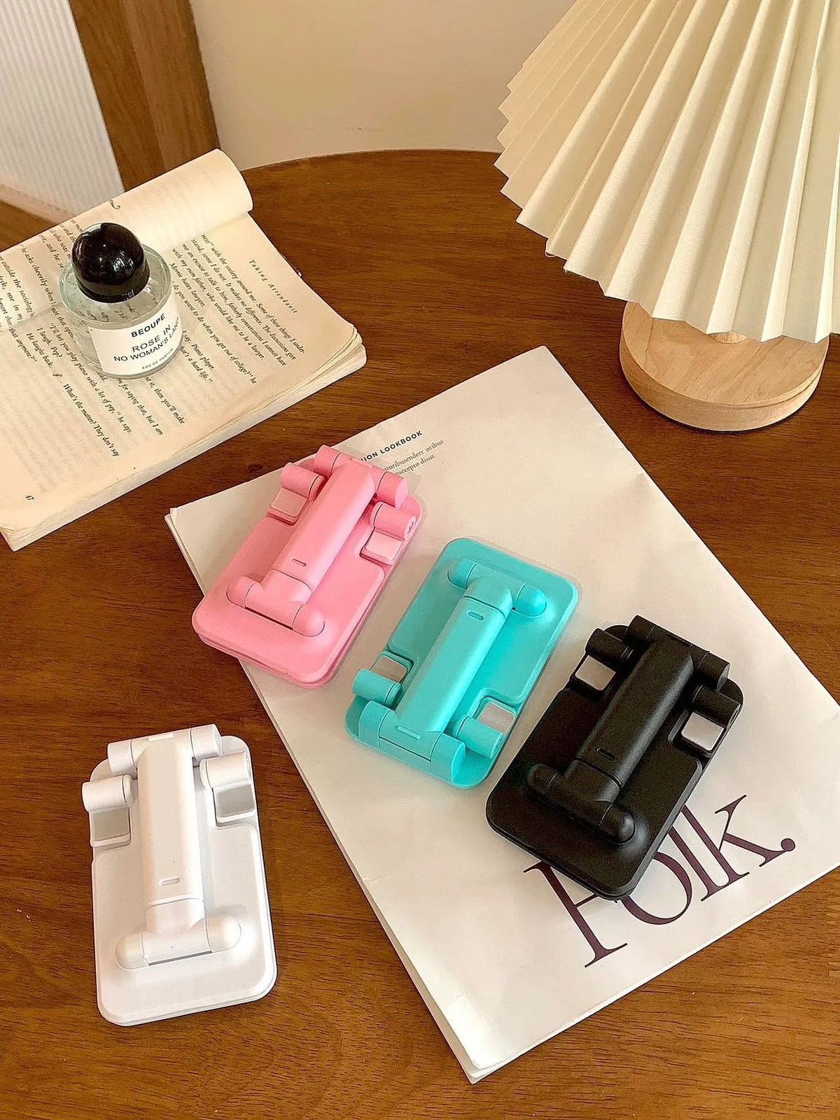 A collection of pink and teal adjustable foldable phone and tablet stands resting on a white paper next to a notebook.