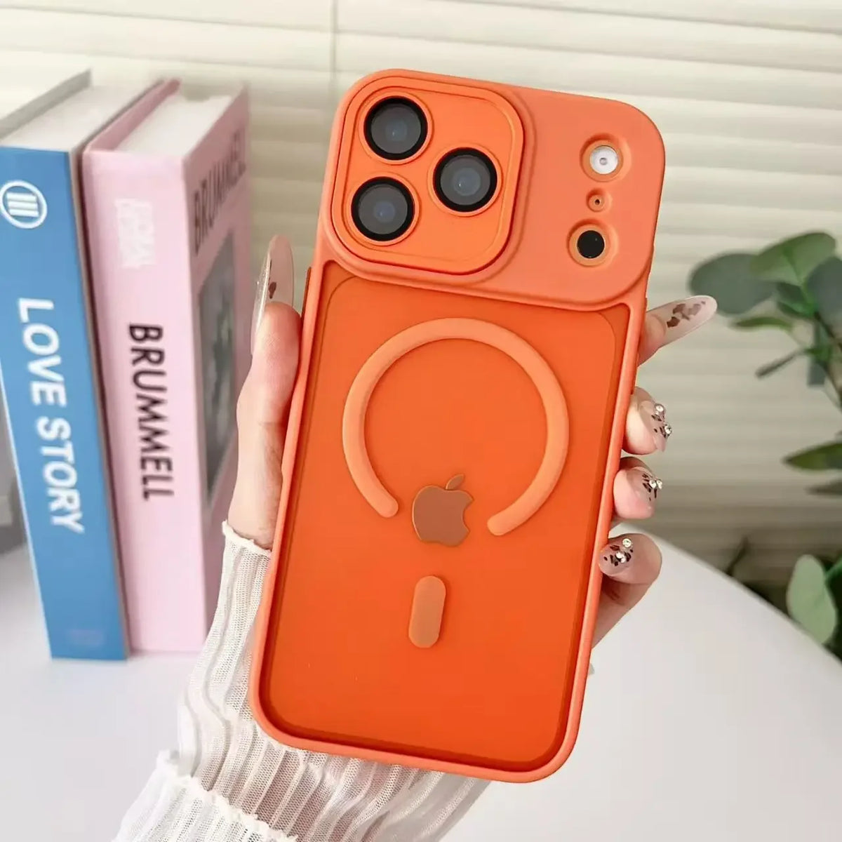 An orange magnetic wireless charging lens protector case for an iPhone is held against a background of books.