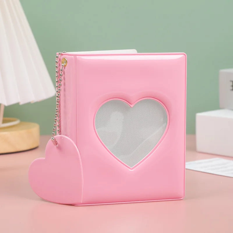 A pink hollow photo card binder with a heart-shaped cutout and a silver chain closure resting on a light pink surface.