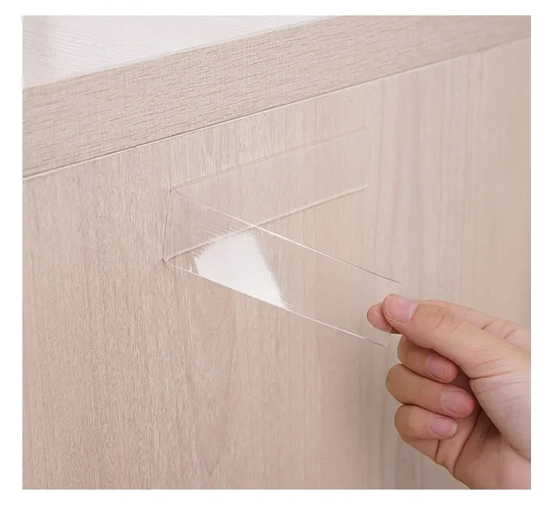 A hand is peeling a piece of clear double-sided nano adhesive tape from a light wood cabinet surface.