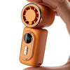 A hand holds an orange portable rechargeable handheld cooling fan with a circular grille on top.