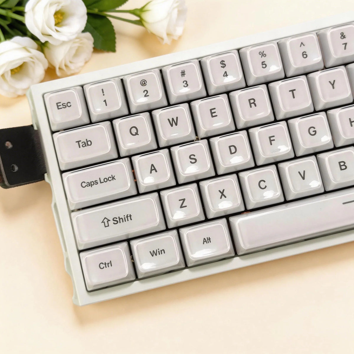A close-up view of a white mechanical keyboard with light gray keycaps and a floral arrangement in the background.