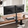 A heavy duty dual monitor desk mount holding two black computer monitors on a wooden desk.