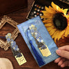 A vintage artistic metal bookmark with a blue tassel is displayed against a dark wooden background next to a sunflower.