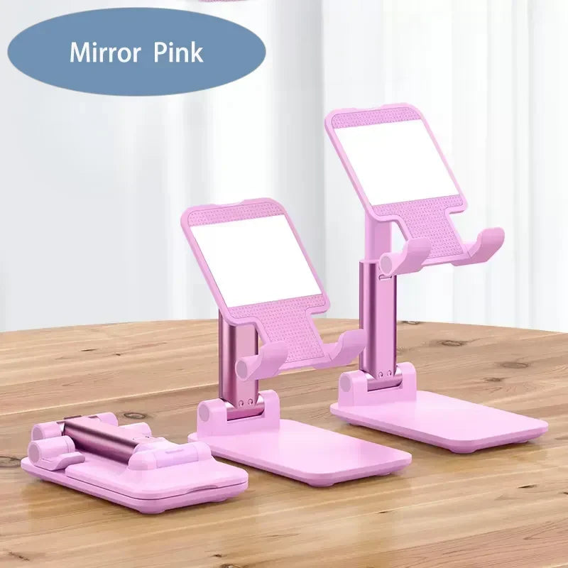 A pink foldable adjustable phone holder stand with a mirror design is shown on a wooden surface.