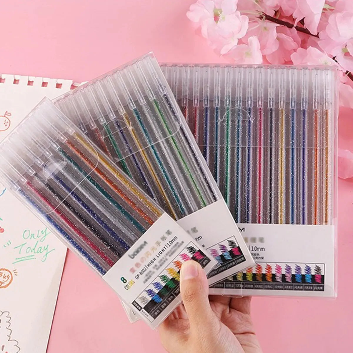 A hand holds a clear plastic case of glitter gel pens with various colors and tips, displayed against a pink background.