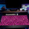 A black gaming mouse pad with a vibrant pink leopard print is displayed on a desk next to a computer monitor.