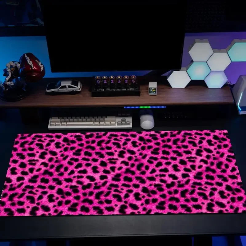 A black gaming mouse pad with a vibrant pink leopard print is displayed on a desk next to a computer monitor.
