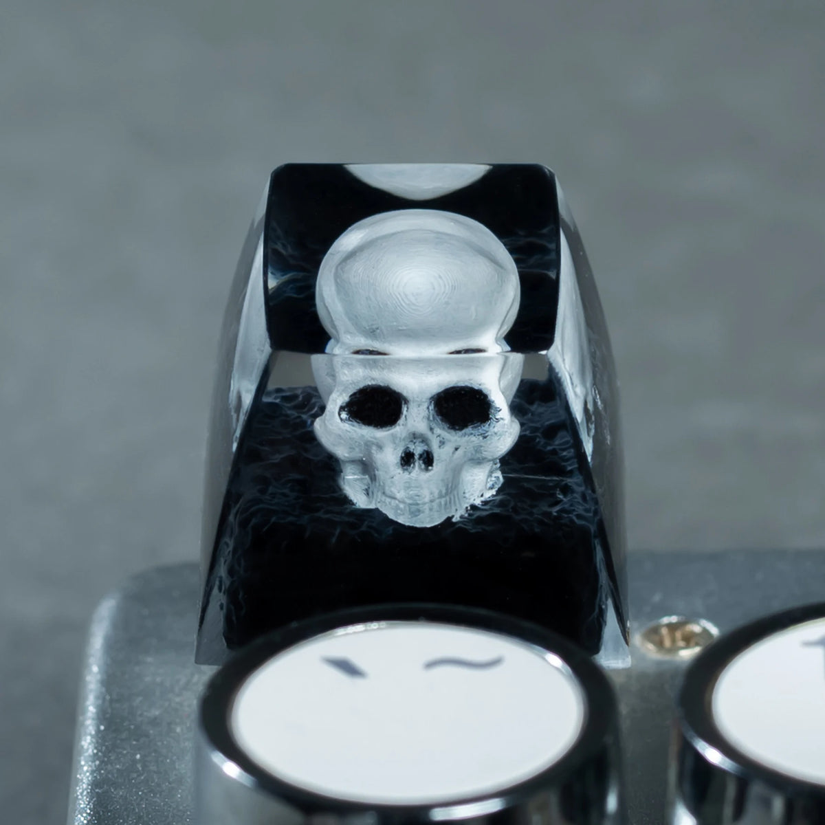 A monochrome skull resin mechanical keycap is shown on a light gray surface.