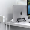 A silver vertical laptop stand holding a MacBook, placed on a white desk next to a white power adapter.