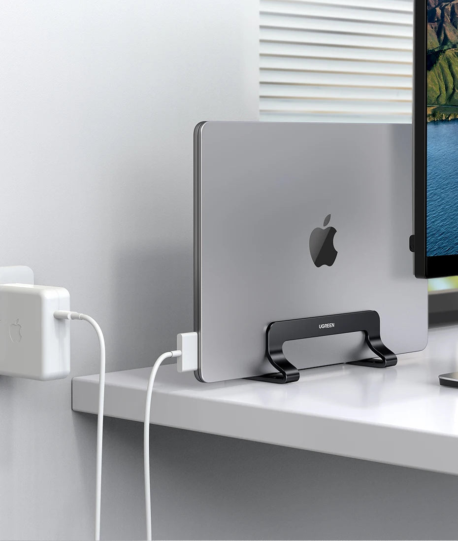 A silver vertical laptop stand holding a MacBook, placed on a white desk next to a white power adapter.