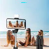 A black Bluetooth fill light selfie stick is shown with a smartphone mounted, set against a beach background.