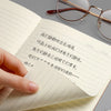 A close-up of an open notebook showing double-sided writing with a pen and eyeglasses on a gray background.