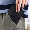 A hand is placing a black retro PU leather pocket notebook into the front pocket of olive green pants, next to a blue and gra