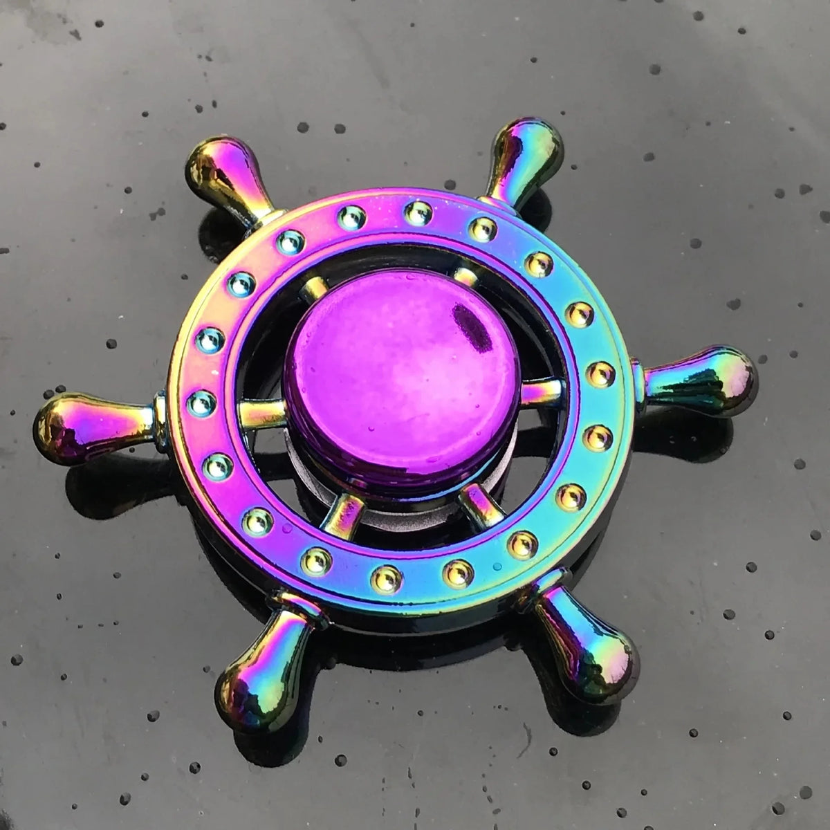 A rainbow metal fidget spinner with a purple center and chrome accents resting on a dark surface.
