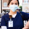 A woman wearing a blue scrubs shirt and a light blue face mask holds an acrylic nurse badge reel.