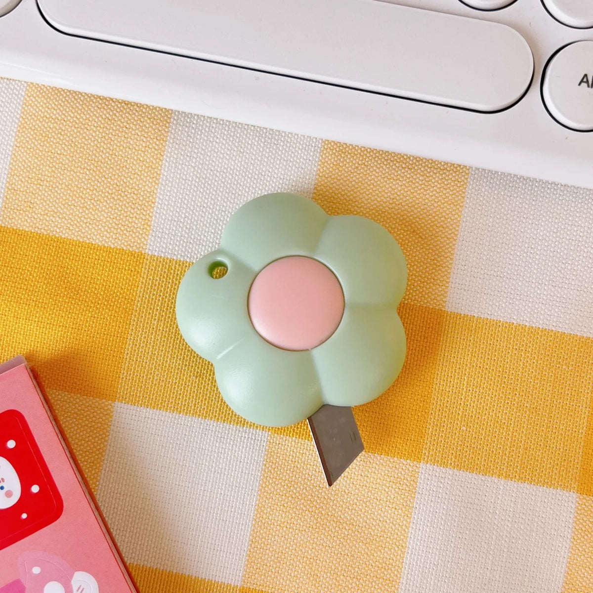 A light green, flower-shaped cat paw mini cutter rests on a yellow and white checkered surface next to a pink box.
