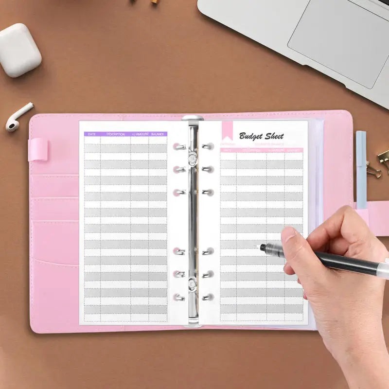 A pink A6 PU leather budget sheet binder with cash envelopes is open on a brown desk next to a laptop.