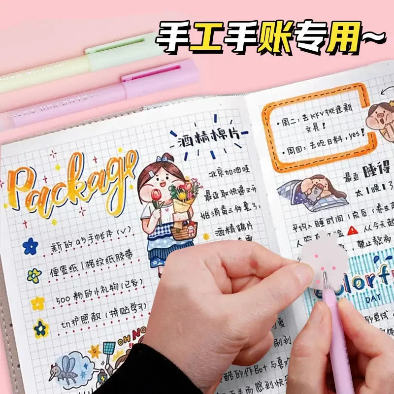 A hand holds a pink glue pen on an open notebook page showing crafting instructions and colorful designs.