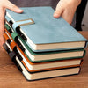 A stack of five A5 retro leather notebooks in shades of blue, green, and brown stacked on a wooden surface.