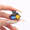 A hand holds a black enamel pin featuring the text you did it and a yellow star with blue accents.