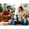 Two men play guitars and sing into a microphone in a bright room with plants.