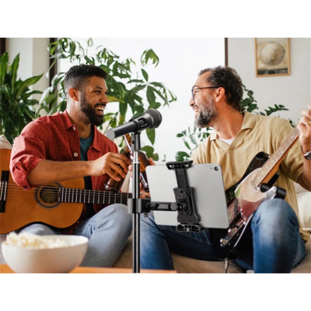 Two men play guitars and sing into a microphone in a bright room with plants.