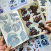 A hand holds an open sticker book filled with fantasy magic themed stickers for laptops and journals