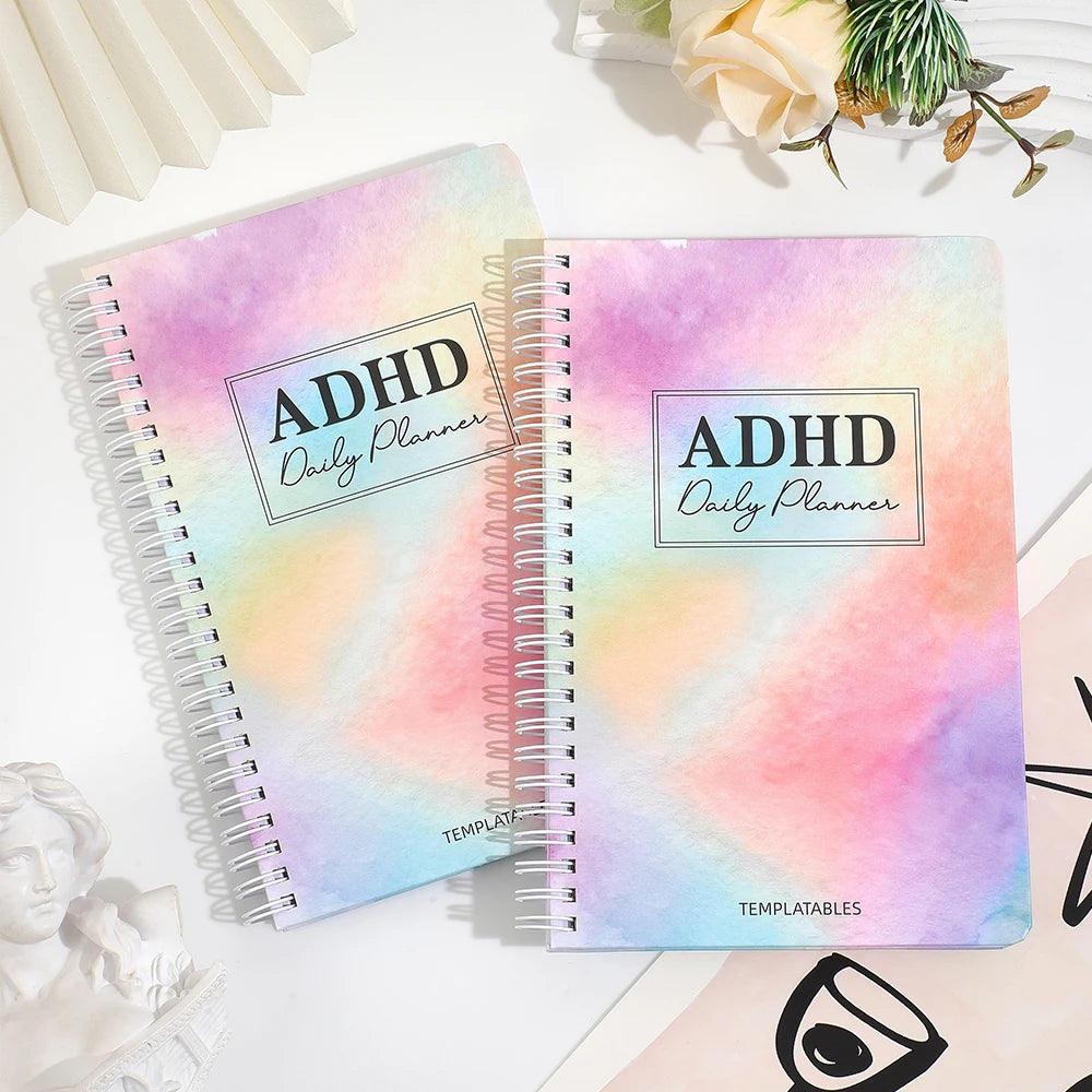 A pink and purple watercolor ADHD Daily Planner notebook with a spiral binding lies on a white surface.