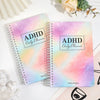 A pink and purple watercolor ADHD Daily Planner notebook with a spiral binding lies on a white surface.