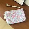 A floral quilted ladies clutch purse with pink and green flowers resting on a wooden surface next to a makeup brush.