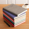 A stack of thick grey notebooks with colored covers resting on a wooden table next to a white cup.