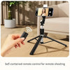 A hand holds a black wireless remote control for a professional extendable selfie stick tripod setup.