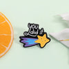 A black enamel pin shaped like a star with the text you did it on it and blue accents
